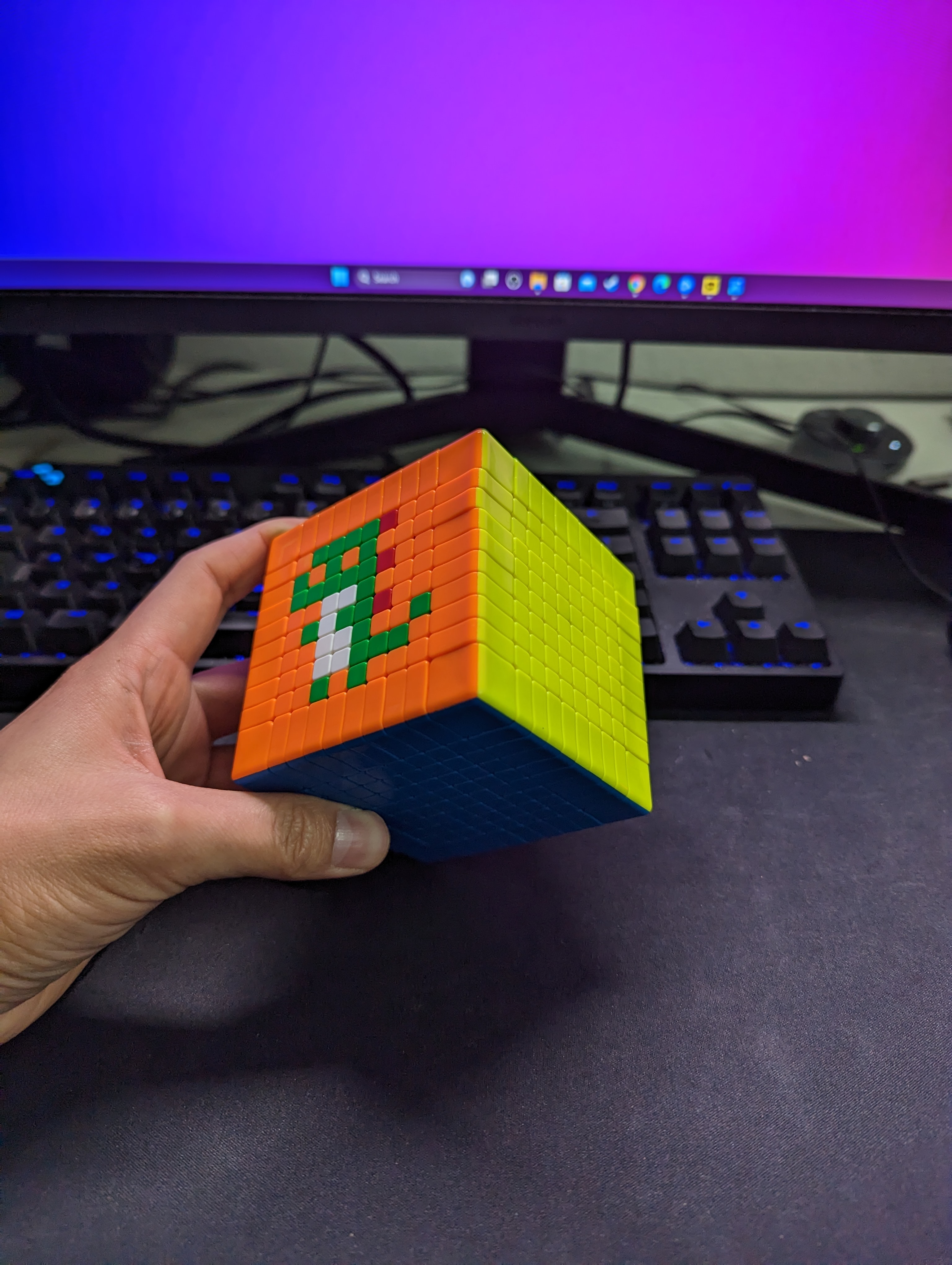 A hand holding the cube to show a green and white pixel figure on the orange front face, with a yellow side and blue underside visible.