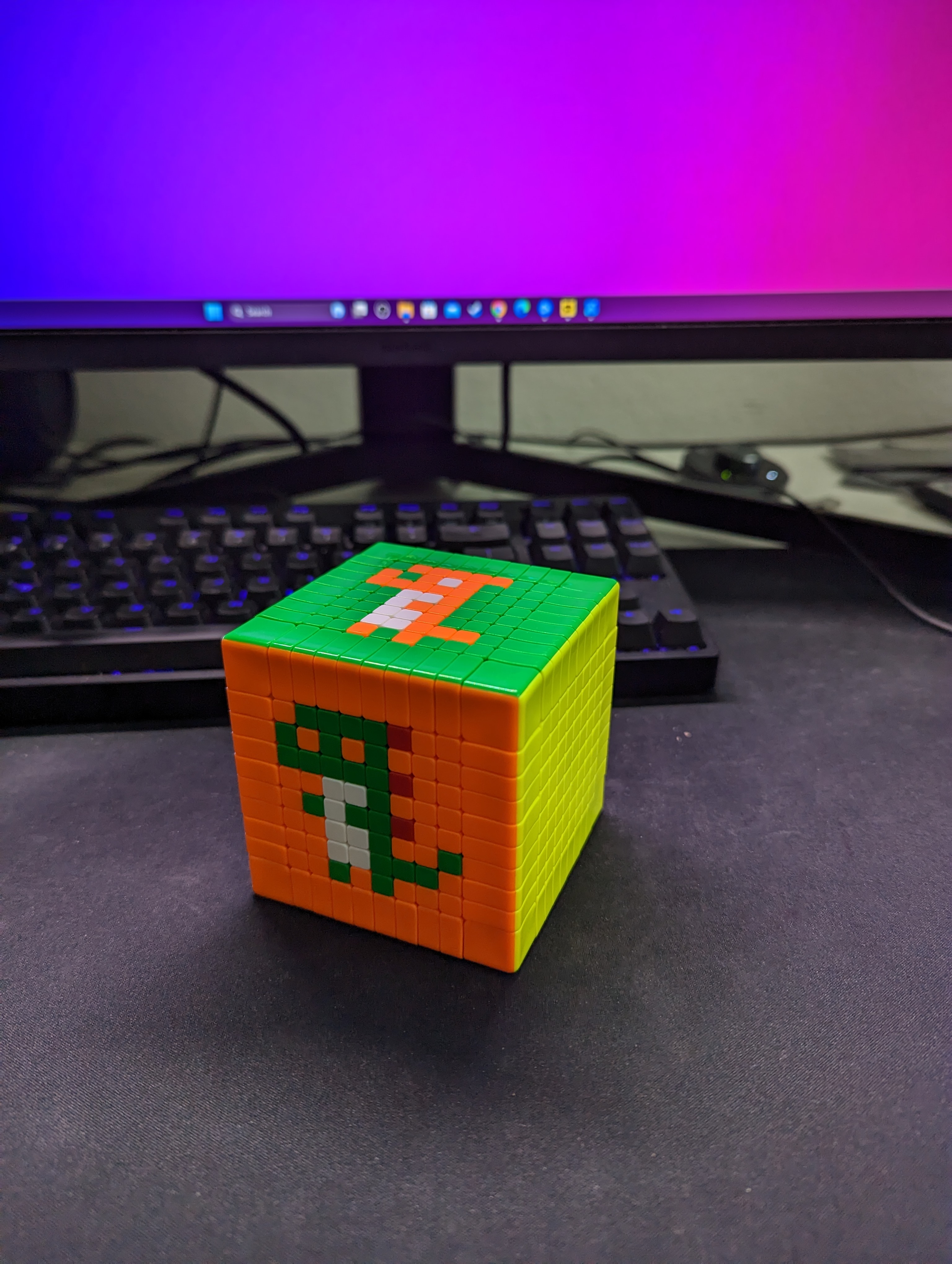 Another view of the cube on a desk, showing the green pixel figure on the orange front face and the orange pixel figure on the green top face.